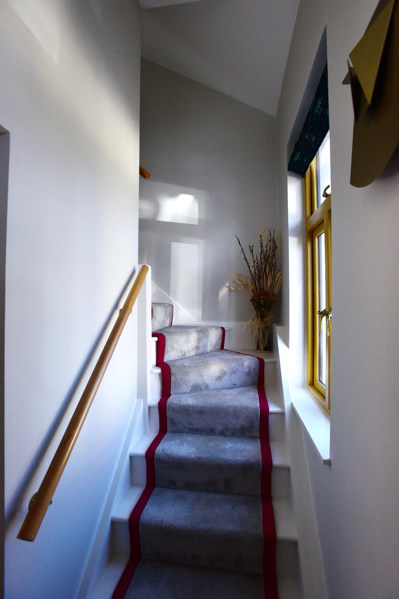 Narrow stairway with grey stair runner bordered in red, beside a yellow-framed window